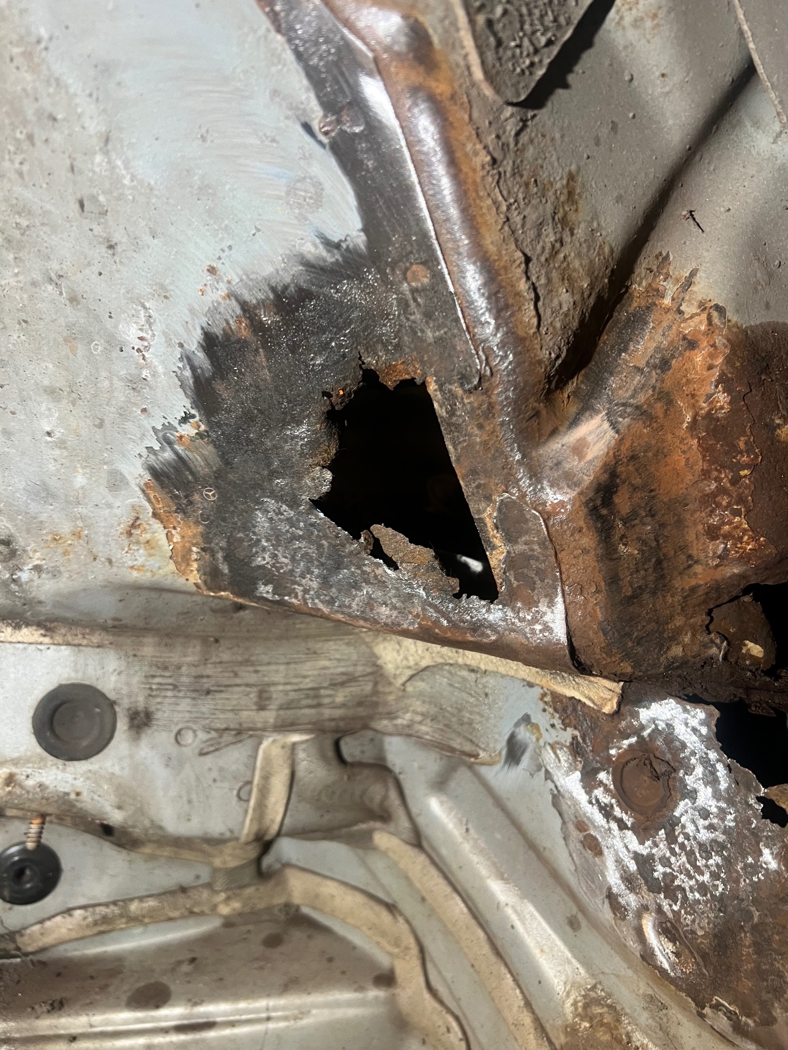 Zbliżenie na skorodowaną blachę samochodową z dziurą, ukazującą zaawansowany proces rdzewienia i uszkodzenia strukturalne. Widoczne ślady wcześniejszych napraw.