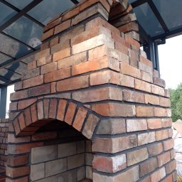 Murowany piec z cegły pod zadaszeniem, widok z boku. Elementy konstrukcyjne z czarną fugą. Widoczne różne odcienie cegieł i fragment otoczenia.