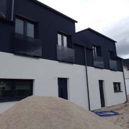 Nowoczesny dom dwukondygnacyjny z ciemną górą i jasnym dołem, widoczny stos piasku, sugerujący prace wykończeniowe. Minimalistyczne balkony z ciemnego szkła.