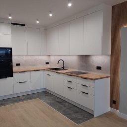 Styl Zakład budowlany Dariusz Stepnowski - Nowoczesna, jasna kuchnia z białymi szafkami, drewnianym blatem i czarną armaturą. Na ścianie dekoracyjne panele z drewna. Podłoga wyłożona płytkami imitującymi kamień.