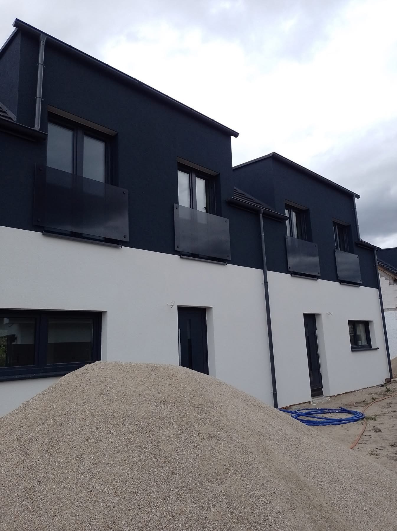 Nowoczesny dom dwukondygnacyjny z ciemną górą i jasnym dołem, widoczny stos piasku, sugerujący prace wykończeniowe. Minimalistyczne balkony z ciemnego szkła.