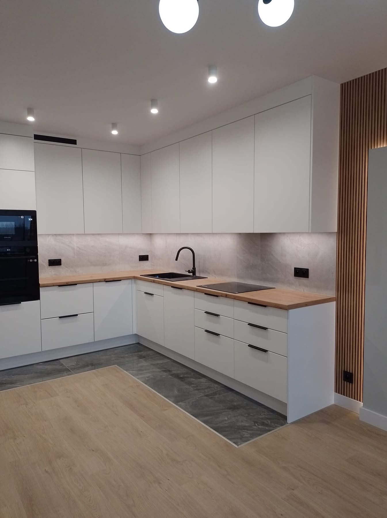 Nowoczesna, jasna kuchnia z białymi szafkami, drewnianym blatem i czarną armaturą. Na ścianie dekoracyjne panele z drewna. Podłoga wyłożona płytkami imitującymi kamień.