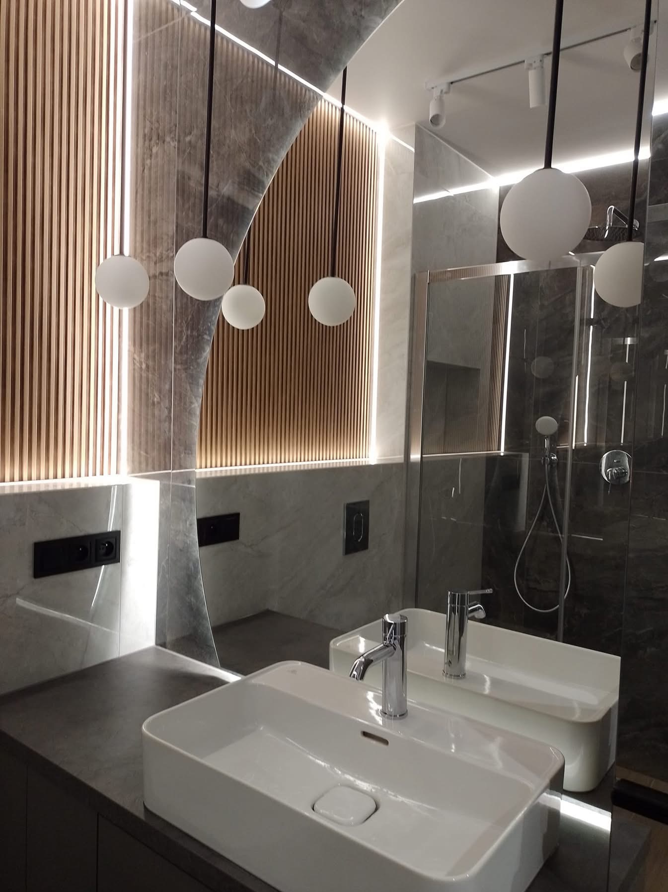 Nowoczesna łazienka z podświetlanymi lamelami, marmurowymi płytami i wiszącymi lampami kulami. Umywalka prostokątna z chromowaną baterią. Widoczne gniazdka i szklana kabina prysznicowa.
