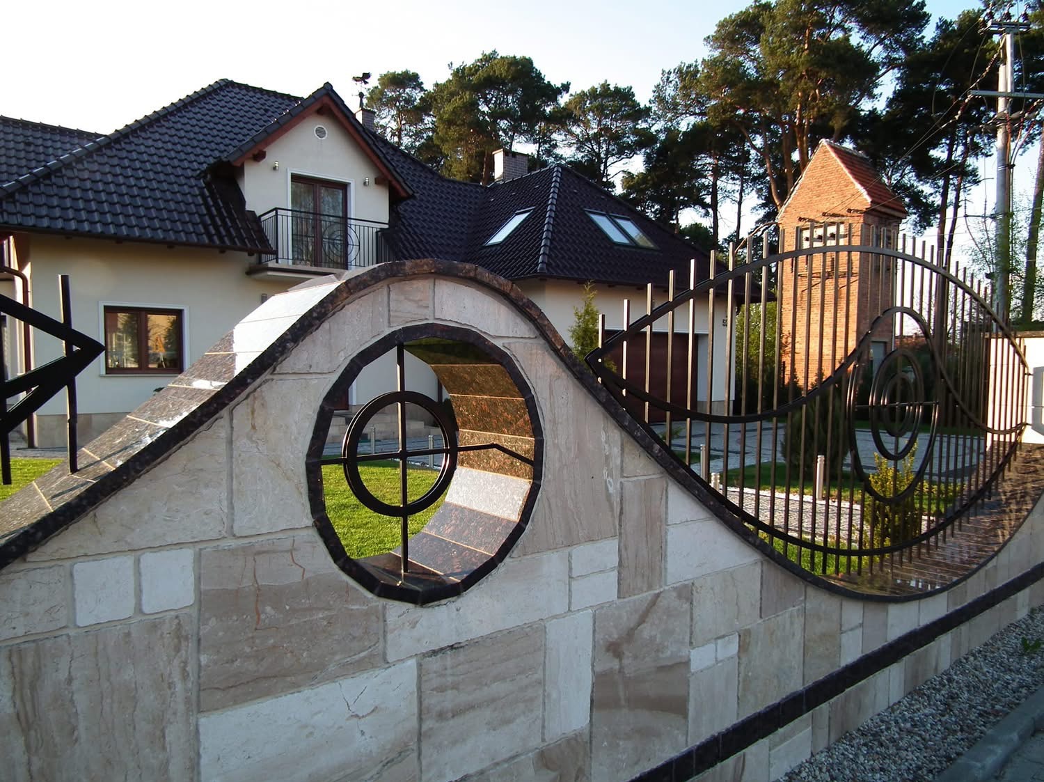 Kamienny mur z dekoracyjnym, metalowym ogrodzeniem i okrągłym prześwitem, przez który widać fragment domu z ciemnym dachem i zielony trawnik.
