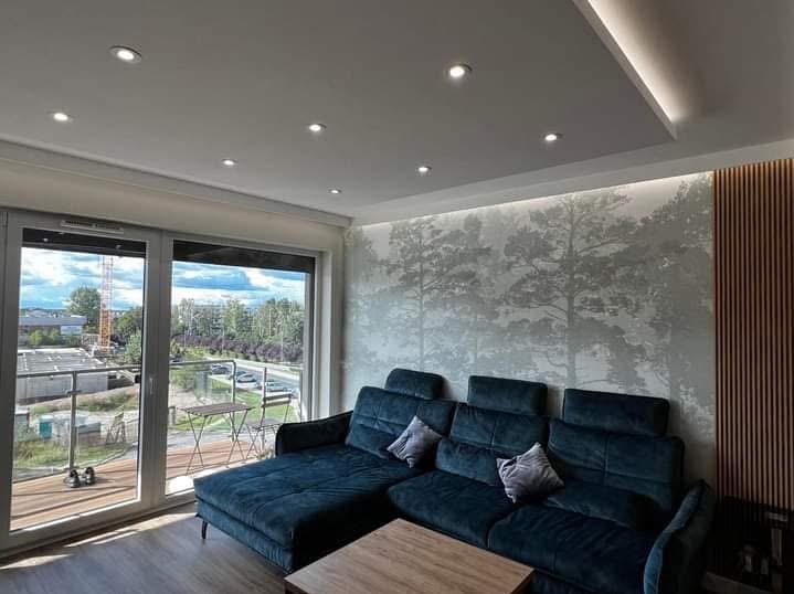 Nowoczesny salon z balkonem: ciemna sofa, ściana z motywem lasu, drewniane panele, sufit z oświetleniem punktowym i listwą LED. Widok na miasto z balkonu.