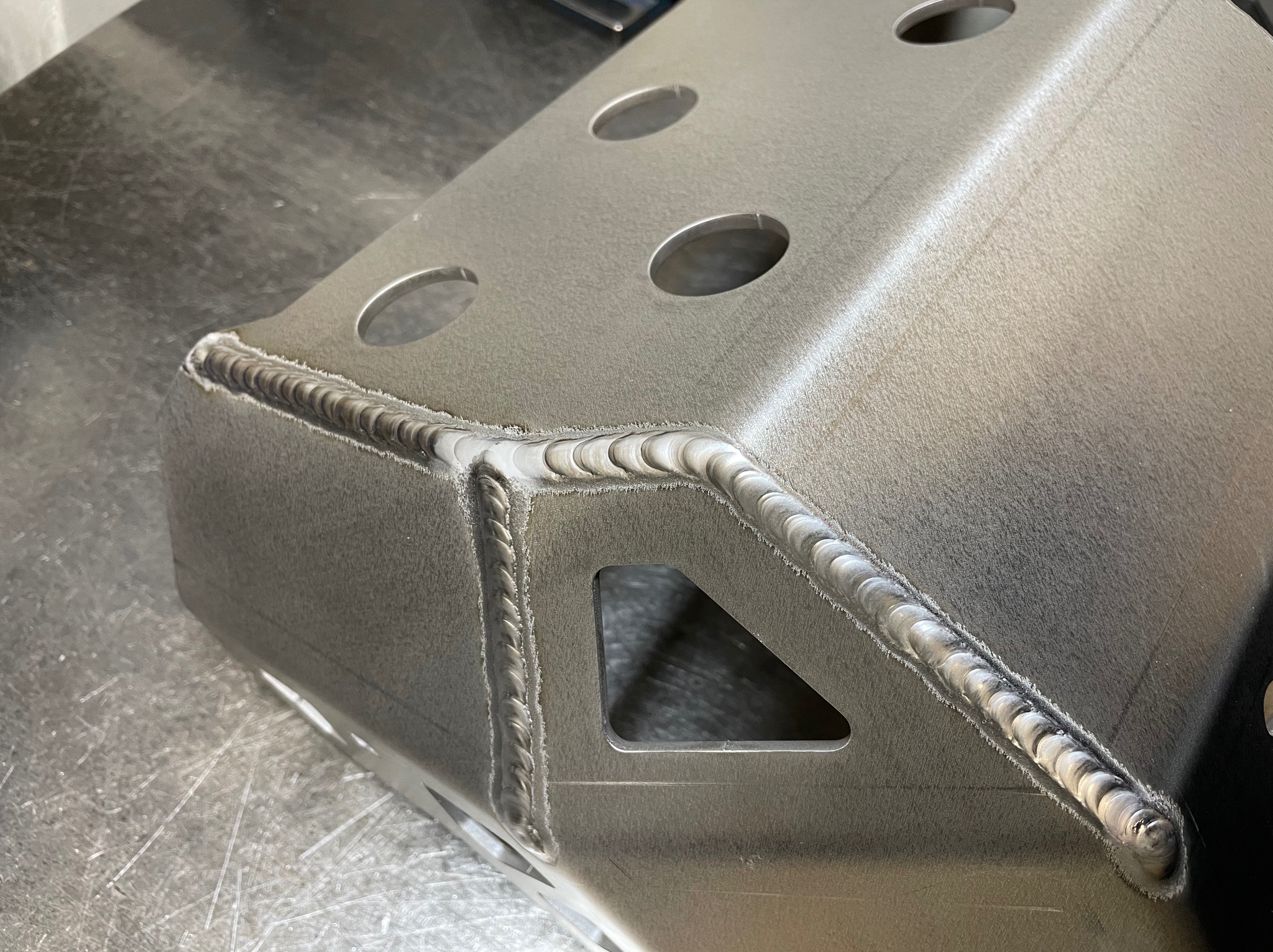 Szlifowany metalowy element z precyzyjnymi spawami i otworami, leżący na metalowym stole. Widoczne ślady obróbki i detale spoin.