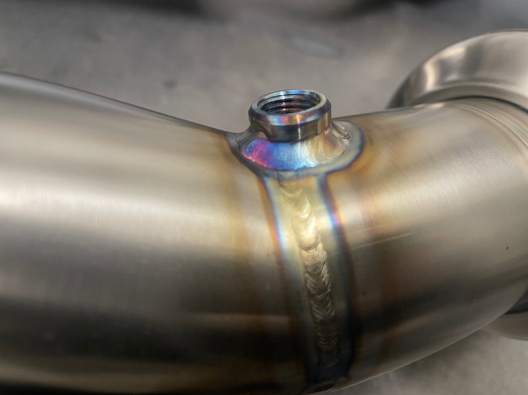Precyzyjne spawy na metalowej rurze z gwintowanym otworem, widoczne tęczowe przebarwienia od ciepła, świadczące o wysokiej jakości wykonania.