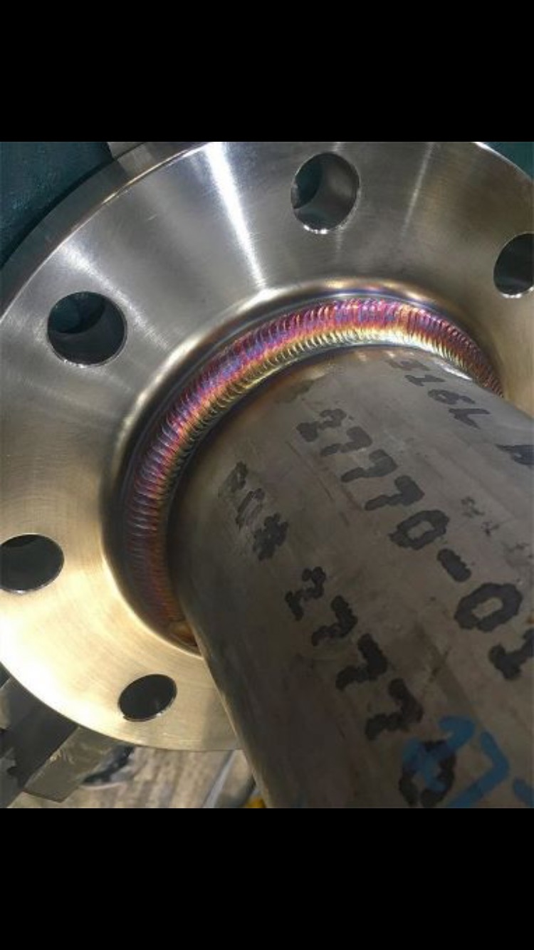 Błyszczące połączenie spawane rury ze stalowym kołnierzem, widoczne tęczowe przebarwienia na spoinie, ciemne napisy na rurze.