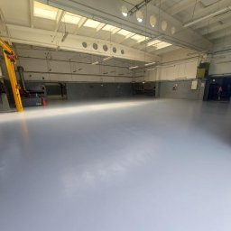 Dawid Schabek Usługi Ogólnobudowlane - Jasnoszara, gładka posadzka w hali przemysłowej z suwnicą i oknami w suficie. Widoczne ślady po obróbce, podkreślające unikalność wykonania.