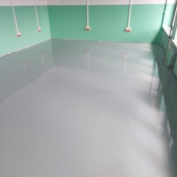 Dawid Schabek Usługi Ogólnobudowlane - Szara, gładka posadzka w pomieszczeniu z zielono-białymi ścianami. Widoczne gniazdka elektryczne na ścianie i odbicie światła w posadzce. Minimalistyczny, przemysłowy styl.