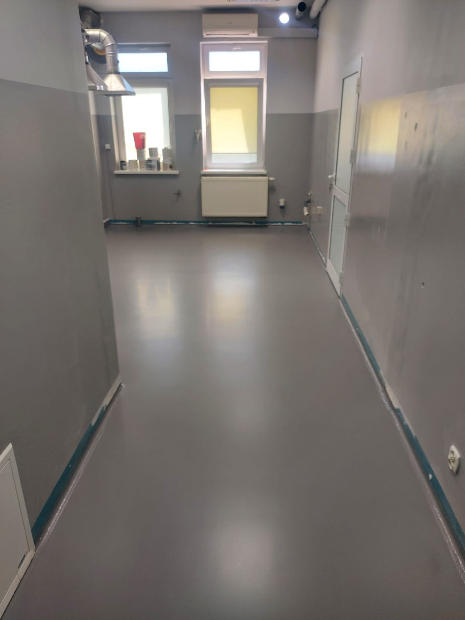 Szara, gładka posadzka w pomieszczeniu z oknami i drzwiami. Widoczne ślady zabezpieczeń przy ścianach. Minimalistyczne wnętrze, jednolite wykończenie podłogi.