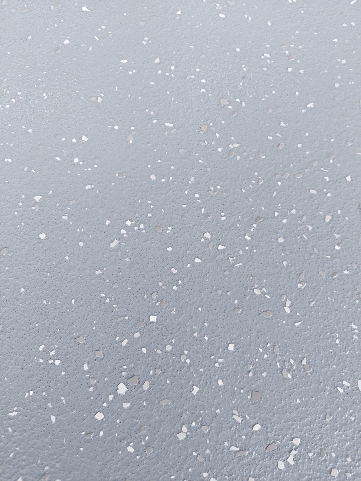Jasnoszara posadzka z drobnymi, nieregularnymi, białymi elementami dekoracyjnymi, tworzącymi unikalną teksturę. Powierzchnia matowa, oświetlenie równomierne.