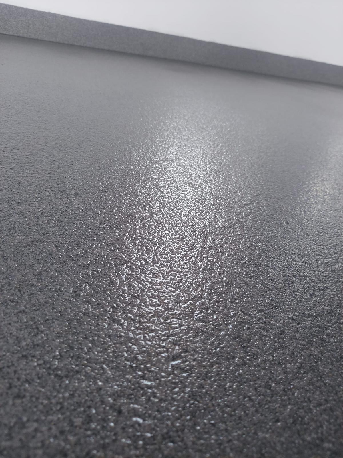 Szara, błyszcząca posadzka z wyraźną fakturą, oświetlona z góry, z widoczną listwą przypodłogową w tle. Delikatne refleksy światła podkreślają nierówności powierzchni.
