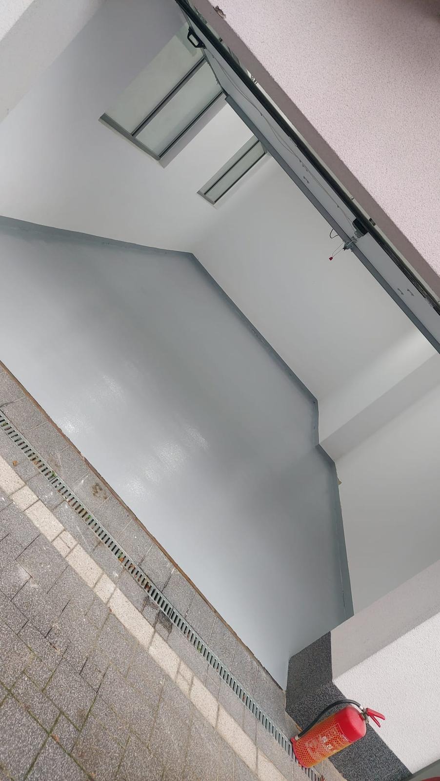 Szara, gładka posadzka w pomieszczeniu z kratką odpływową przy ścianie i gaśnicą w rogu. Widoczne okno dachowe i fragment sufitu.