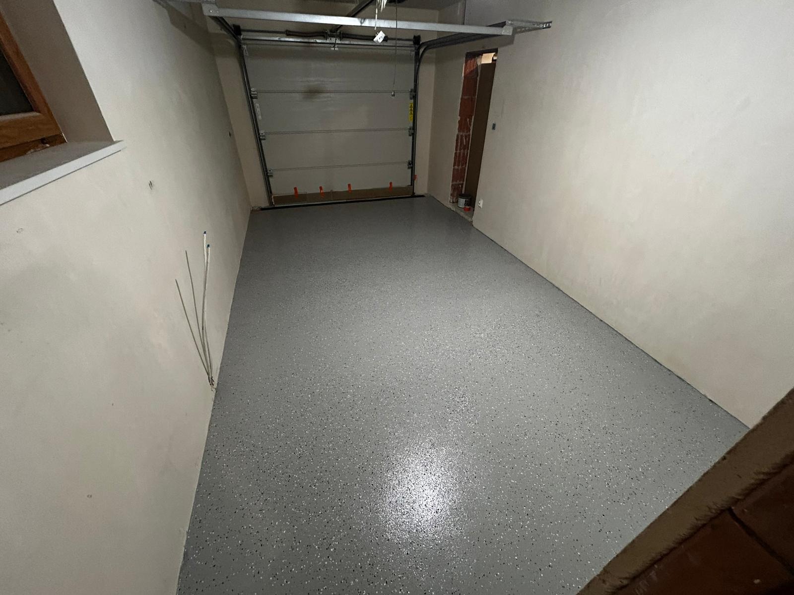 Szara posadzka z drobnymi, ciemnymi plamkami w garażu z bramą segmentową. Świeżo wylana, gładka powierzchnia. Widoczne ściany i fragment okna.