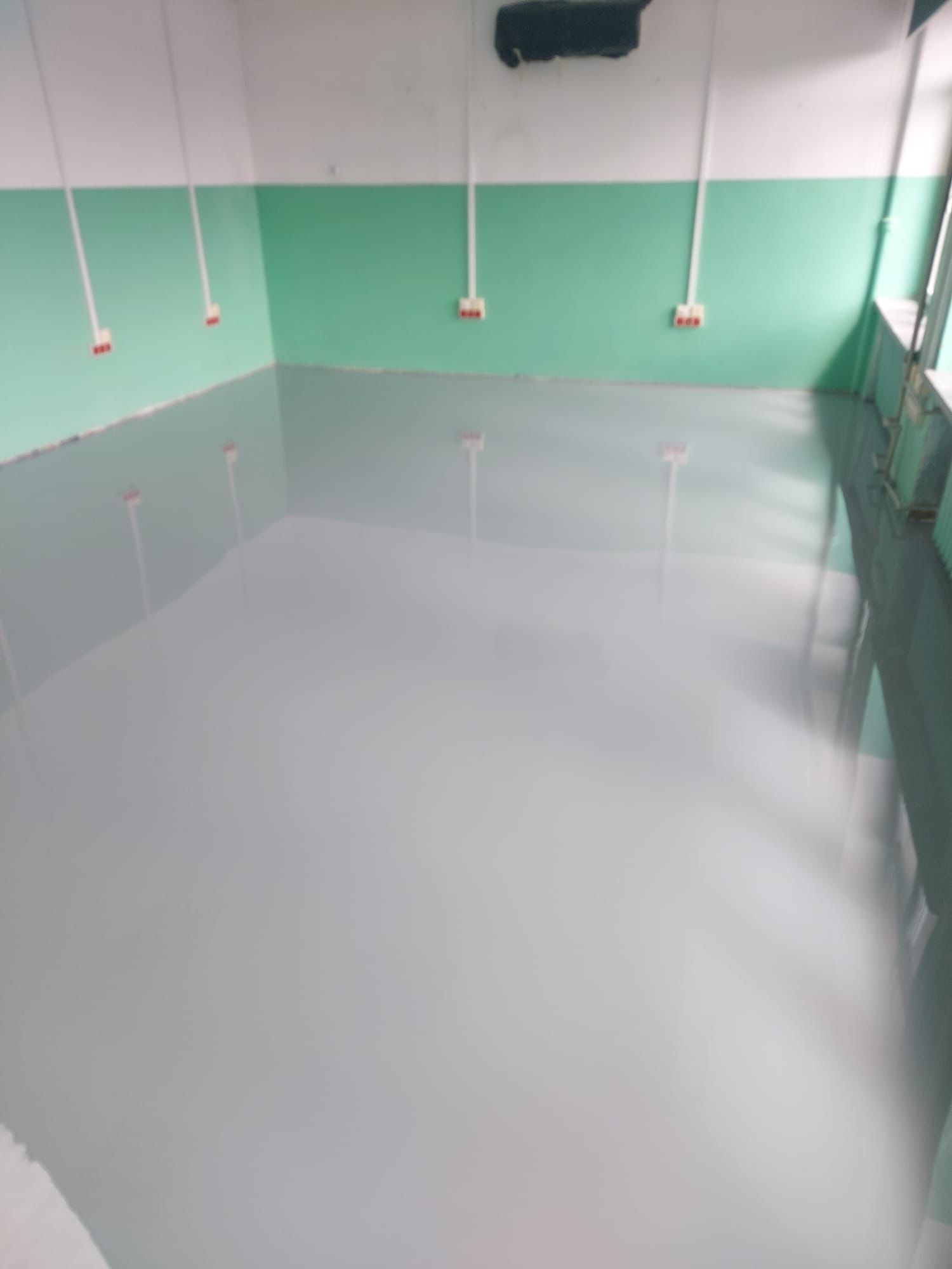 Szara, gładka posadzka w pomieszczeniu z zielono-białymi ścianami. Widoczne gniazdka elektryczne na ścianie i odbicie światła w posadzce. Minimalistyczny, przemysłowy styl.