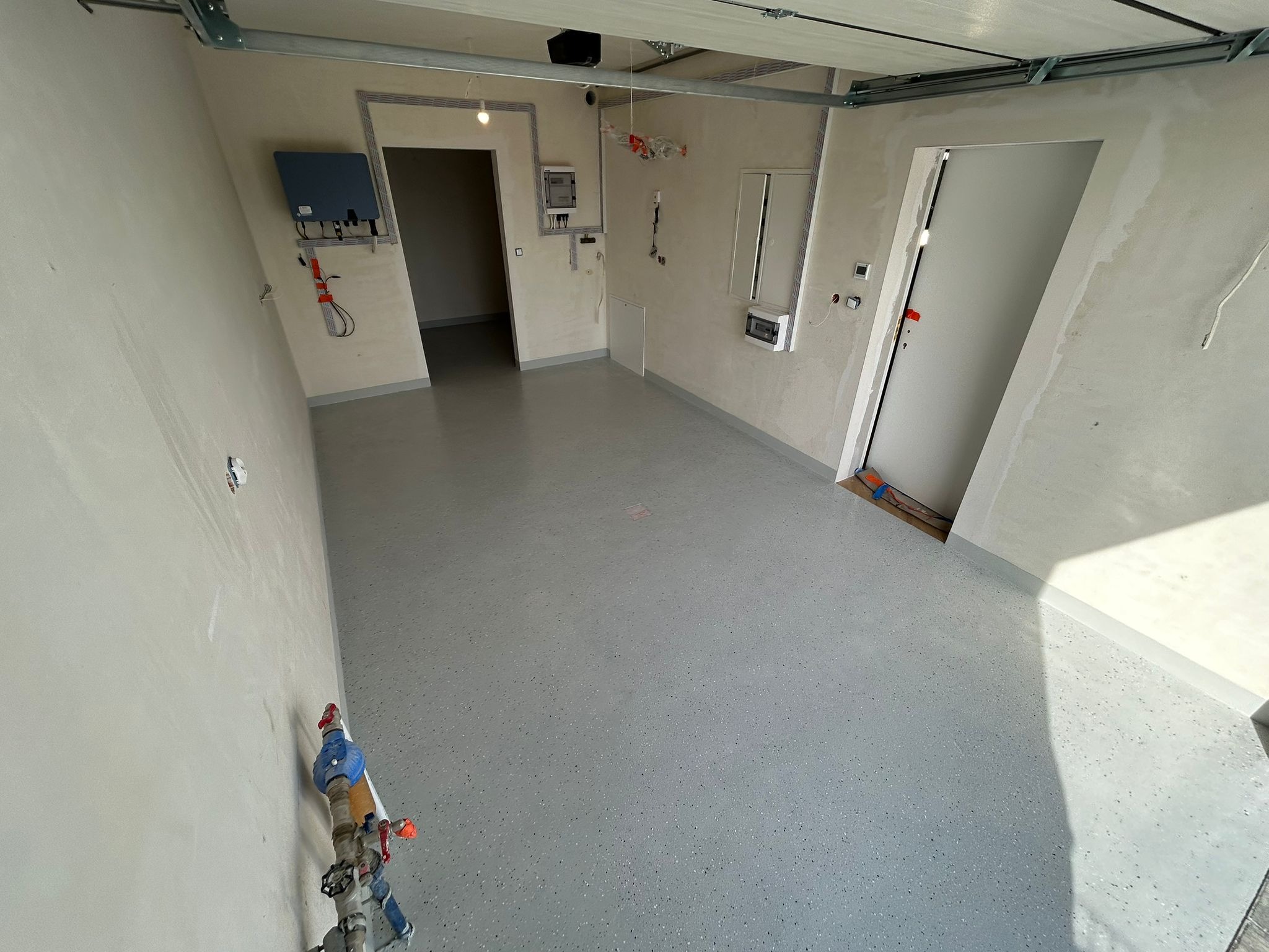 Szara posadzka z drobnymi plamkami w garażu. Widoczne instalacje na ścianach i drzwi. Świeżo wylana, gładka powierzchnia. Minimalistyczny, surowy styl wykończenia.