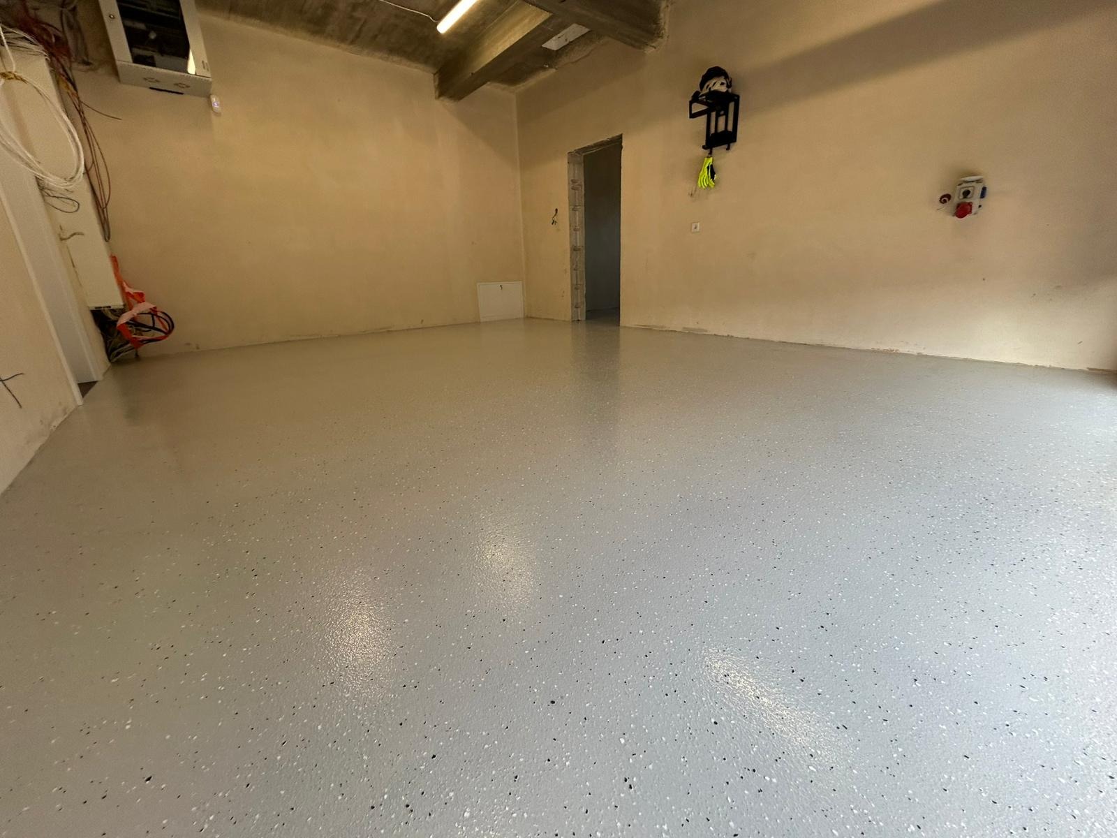 Jasnoszara posadzka żywiczna z drobnymi, ciemnymi inkluzjami w pustym pomieszczeniu z beżowymi ścianami i betonowym sufitem. Widoczne instalacje elektryczne.