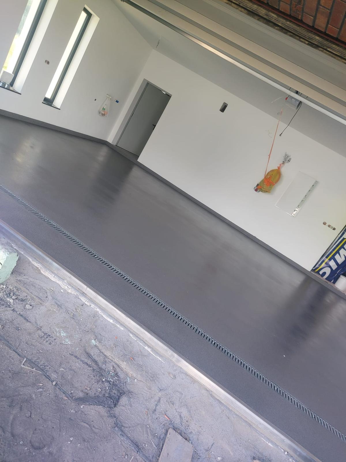 Szara, gładka posadzka w garażu/pomieszczeniu gospodarczym z odpływem liniowym przy bramie. Świeżo wylana, lśniąca powierzchnia. Białe ściany, okna w tle.