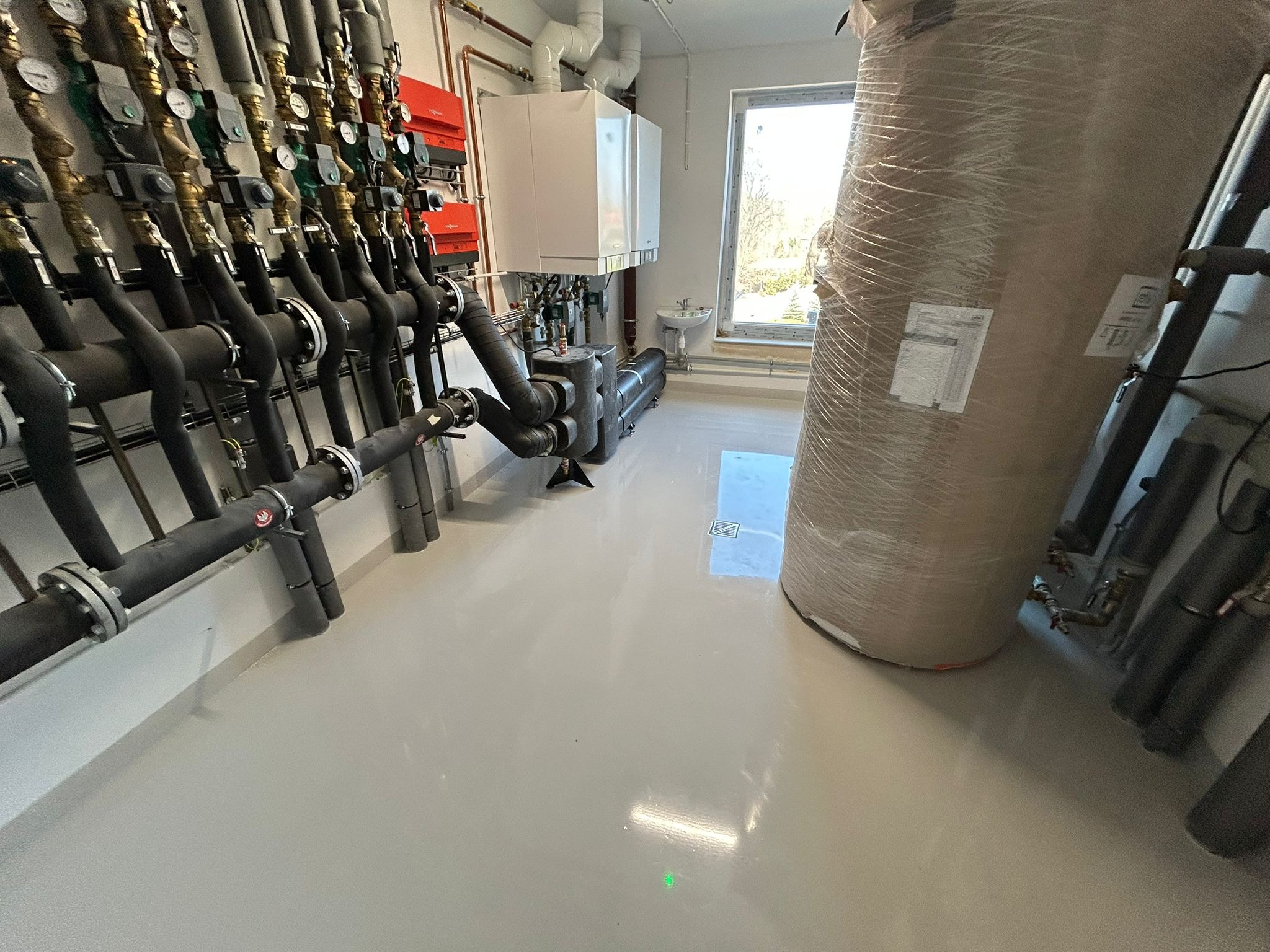 Jasna, gładka posadzka w kotłowni z instalacją grzewczą. Widoczne rury, zawory i zbiornik ciepłej wody. Minimalistyczny styl, czyste linie, nowoczesne wykończenie.