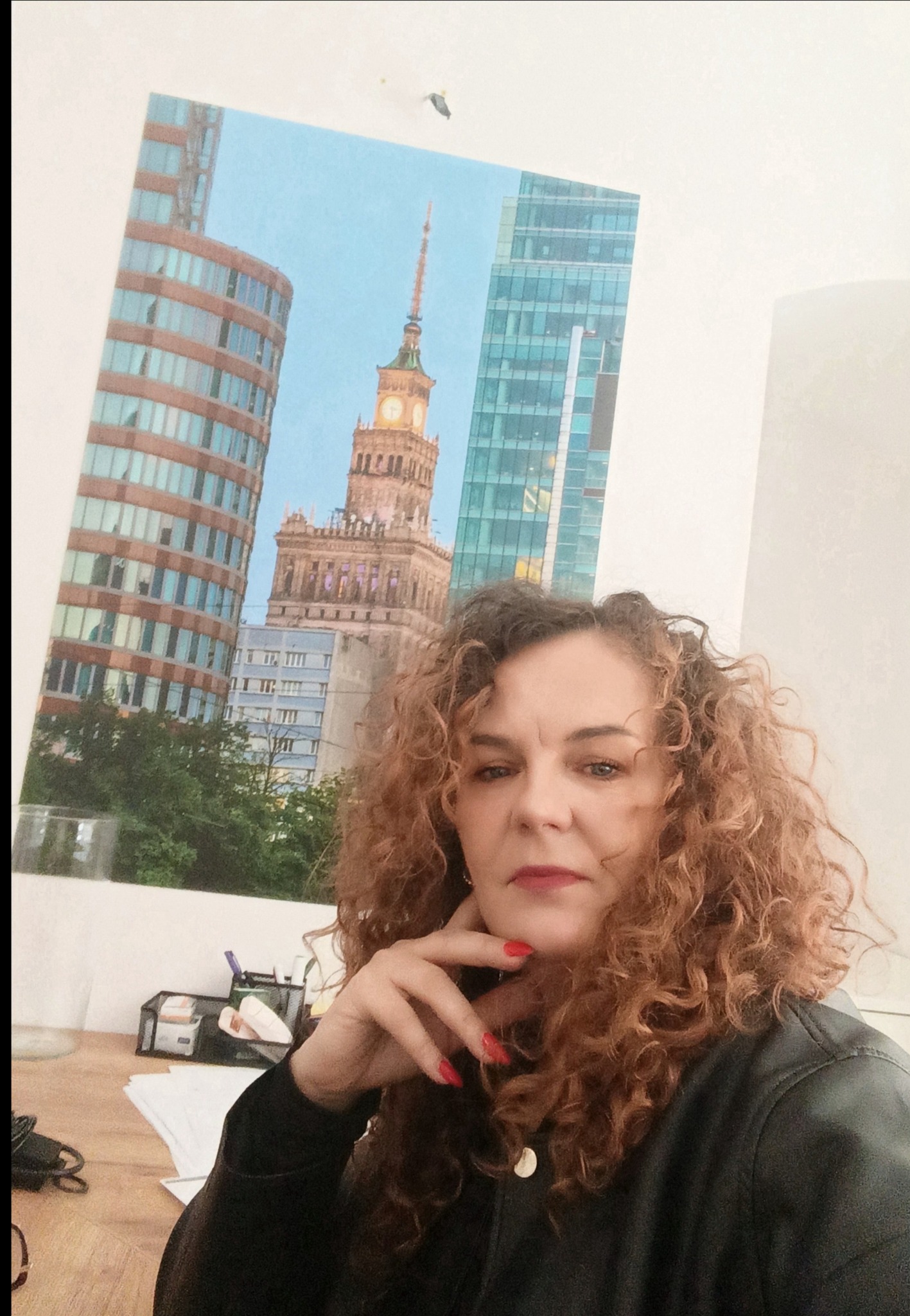 Kobieta z kręconymi włosami pozuje na tle plakatu z Pałacem Kultury i Nauki w Warszawie. Uwagę przykuwają czerwone paznokcie i skórzana kurtka.