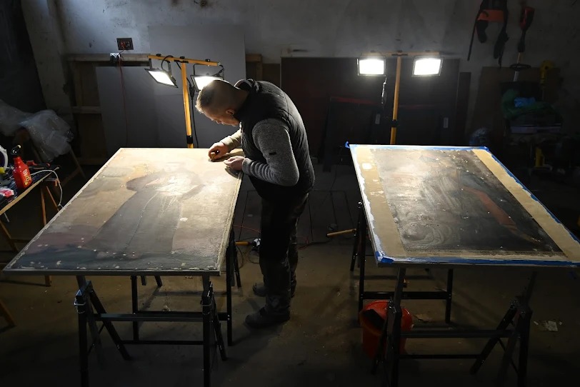 Prace konserwatorskie: mężczyzna delikatnie czyści zabytkowy obraz na stole warsztatowym, drugi obraz obok, oświetlenie halogenowe, surowe wnętrze pracowni.