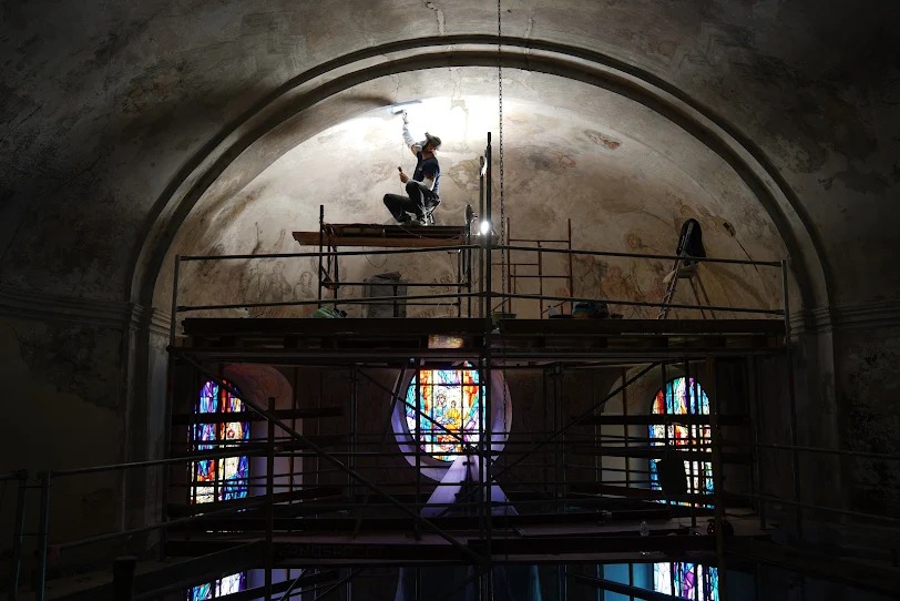 Prace konserwatorskie w kościele: mężczyzna na rusztowaniu odnawia malowidła na sklepieniu, w tle witraże. Praca z użyciem narzędzi i oświetlenia.