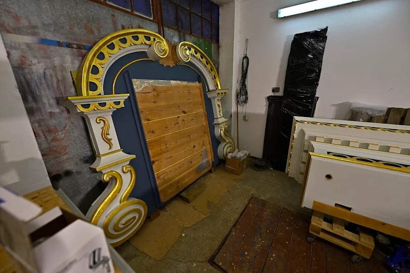 Elementy dekoracyjne z drewna, złocone, w trakcie renowacji. Widoczne fragmenty konstrukcji i zabezpieczone elementy. Warsztat z oknami w tle.