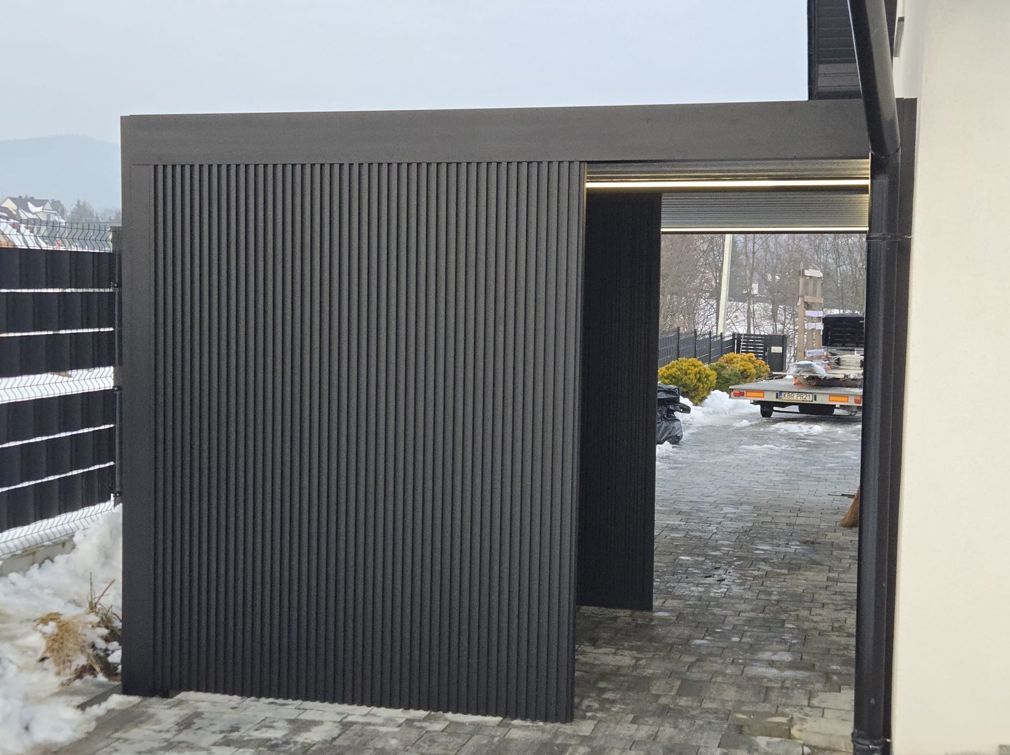 Nowoczesna, grafitowa konstrukcja z pionowych paneli, prawdopodobnie zadaszenie tarasu, z oświetleniem LED, widoczna z zewnątrz na tle zimowego krajobrazu.