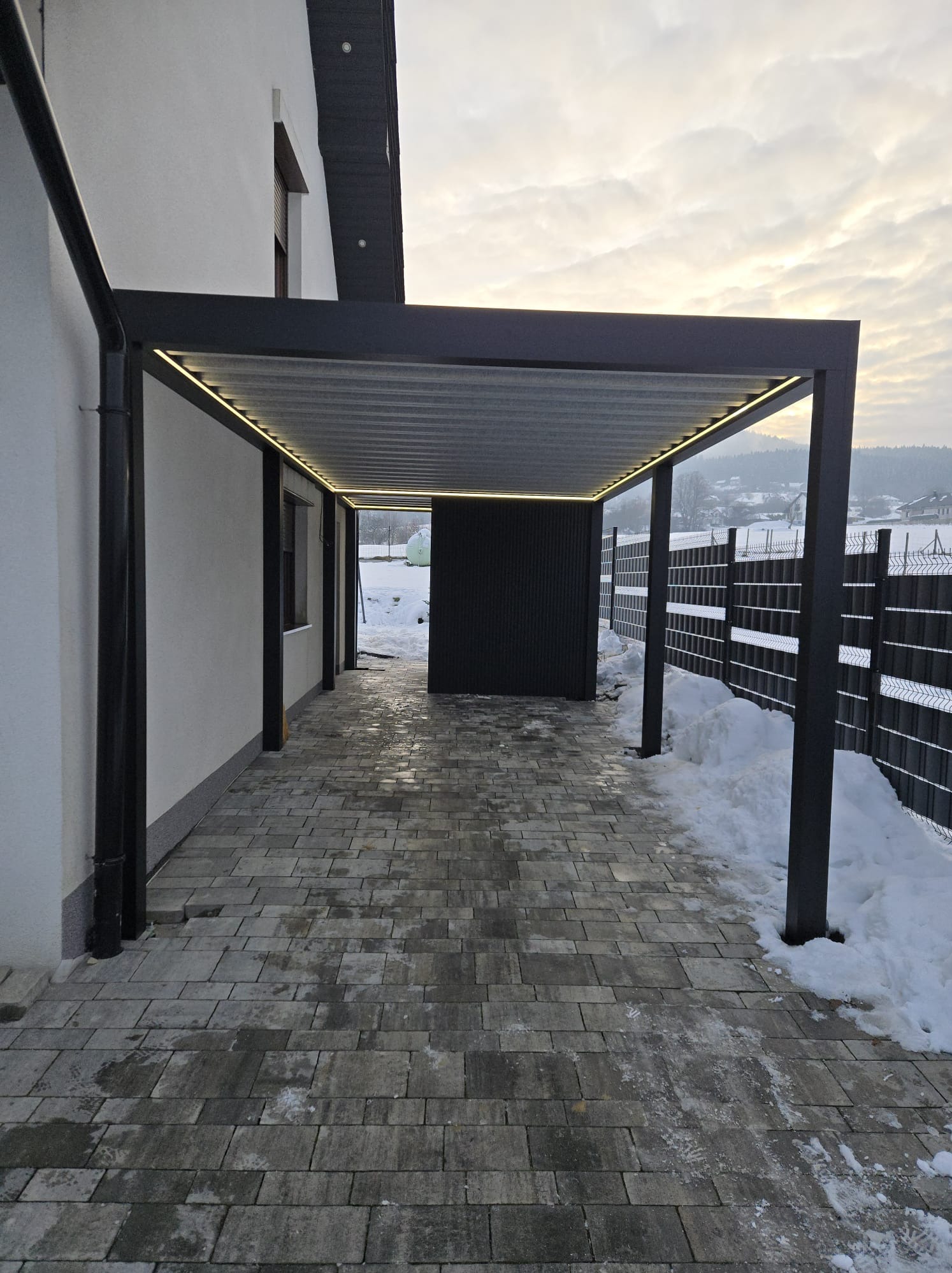 Nowoczesna, grafitowa konstrukcja z oświetleniem LED, przylegająca do budynku. Widoczny fragment posesji w zimowej scenerii i ogrodzenie panelowe.