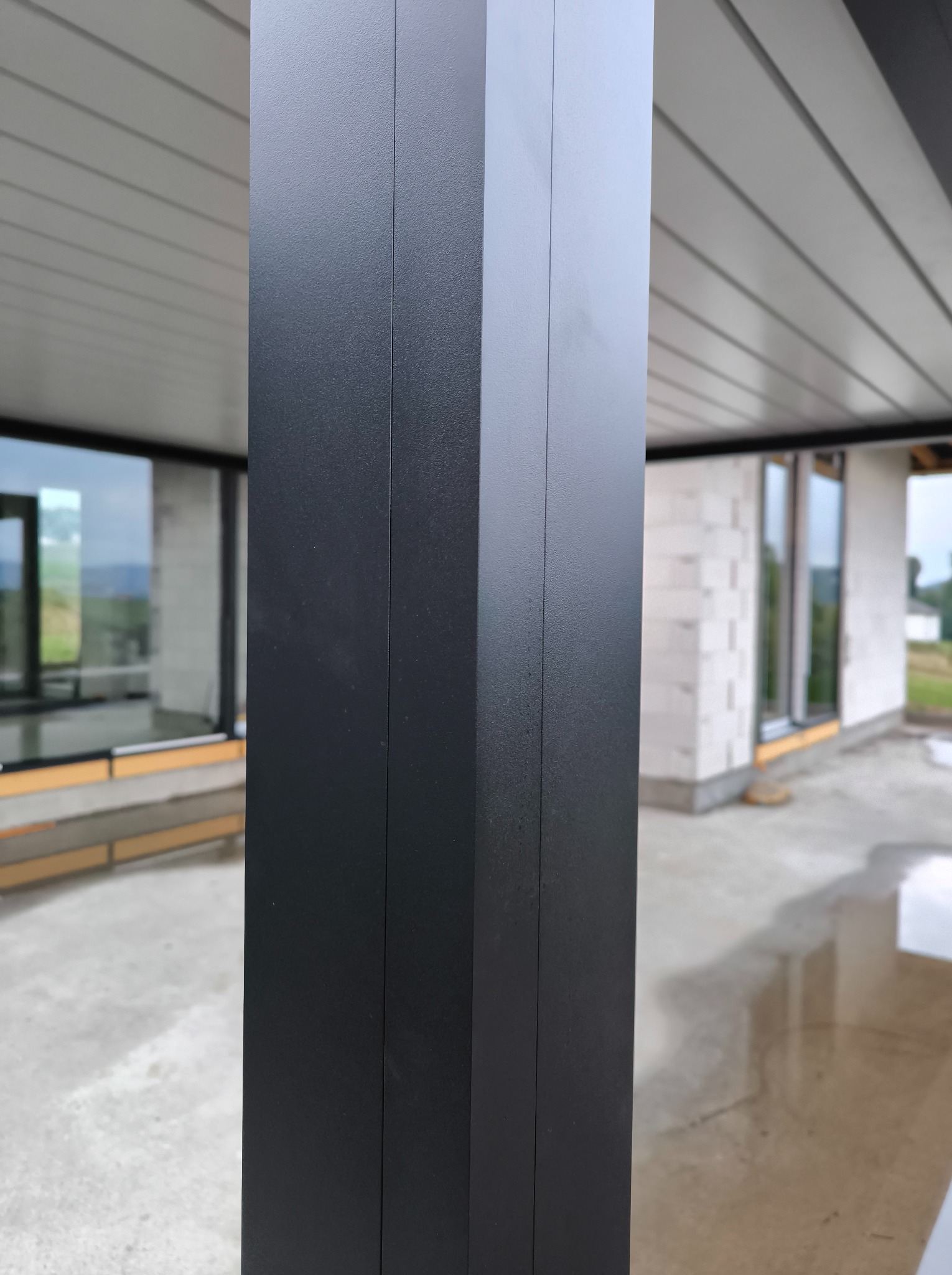 Fragment konstrukcji z czarnego metalu, prawdopodobnie słup podtrzymujący zadaszenie tarasu. W tle widoczne okno i ściana budynku w trakcie budowy.