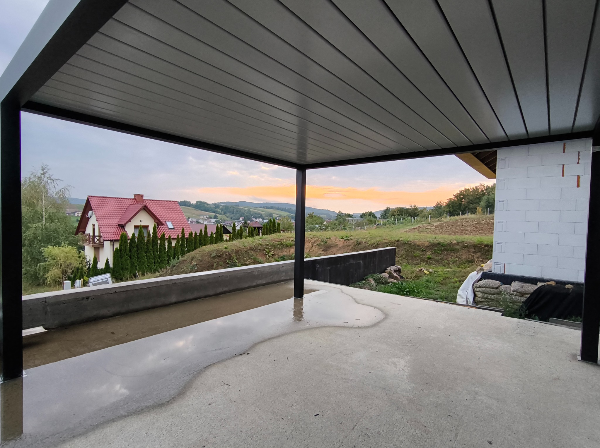 Nowoczesna, grafitowa konstrukcja zadaszenia tarasu z widokiem na dom z czerwoną dachówką i malowniczy krajobraz o zachodzie słońca. Betonowa podłoga w stanie surowym.
