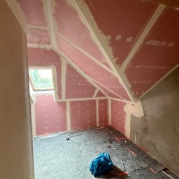 Poddasze z oknem dachowym w trakcie prac wykończeniowych: różowe płyty GK na skosach i ścianach, widoczne łączenia, częściowo położona gładź, zabezpieczona podłoga.