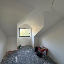 Poddasze po malowaniu na biało, widoczne okno w szczycie, zabezpieczona podłoga, narzędzia i stół z akcesoriami malarskimi. Świeżo pomalowane ściany i sufit.