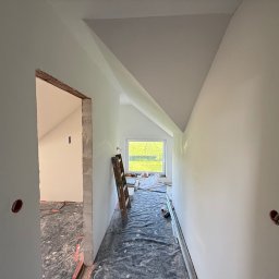 Białe ściany poddasza po malowaniu, widok z korytarza. Widoczne okno, drabina oparta o ścianę i zabezpieczona podłoga. Otwór w ścianie z instalacją elektryczną.