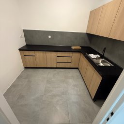 Martin Zawada - Nowoczesna kuchnia z drewnianymi szafkami i czarnym blatem, szare płytki na podłodze i ścianie. Ujęcie z perspektywy wejścia, widoczny fragment ościeżnicy.