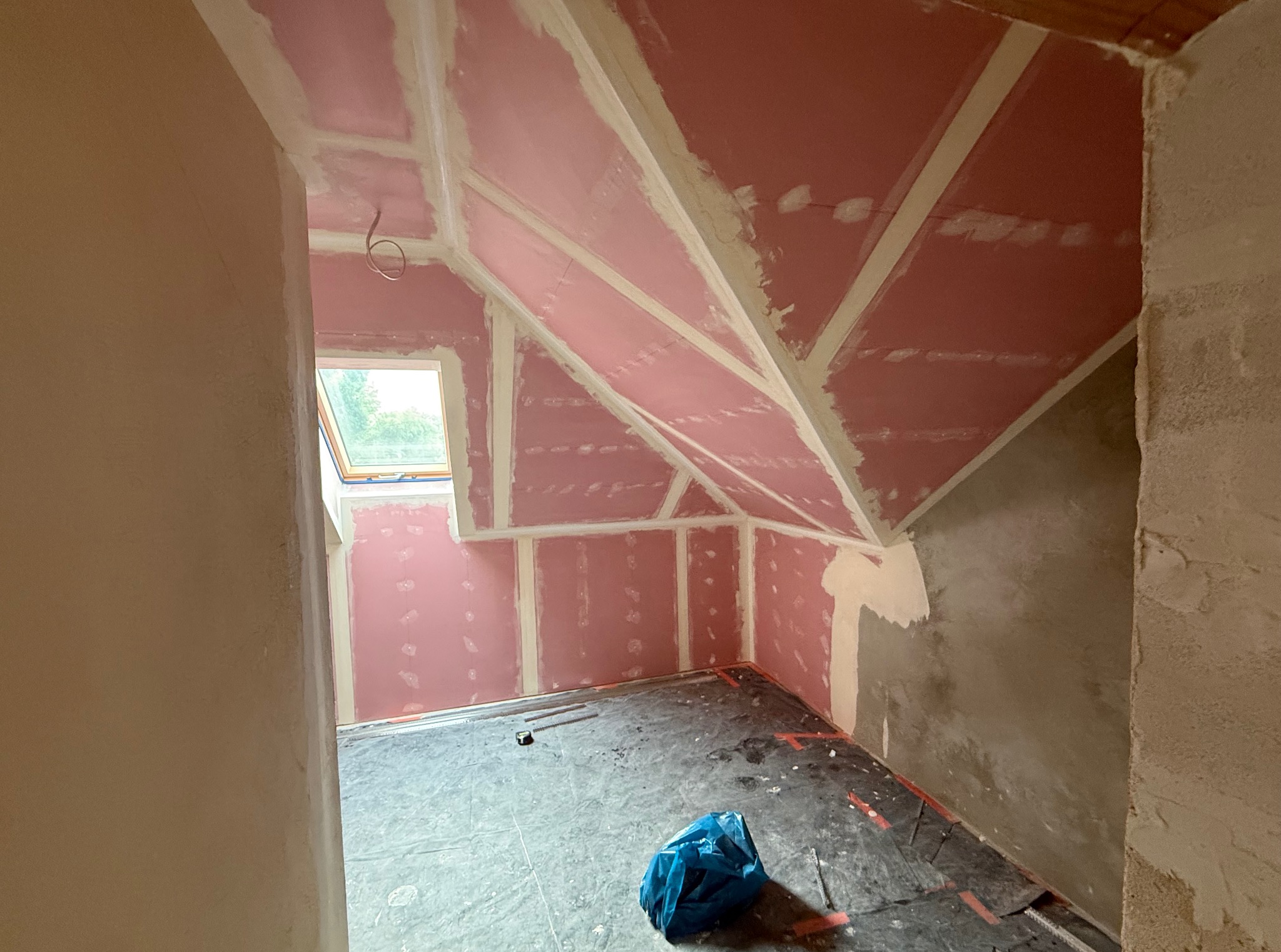Poddasze z oknem dachowym w trakcie prac wykończeniowych: różowe płyty GK na skosach i ścianach, widoczne łączenia, częściowo położona gładź, zabezpieczona podłoga.