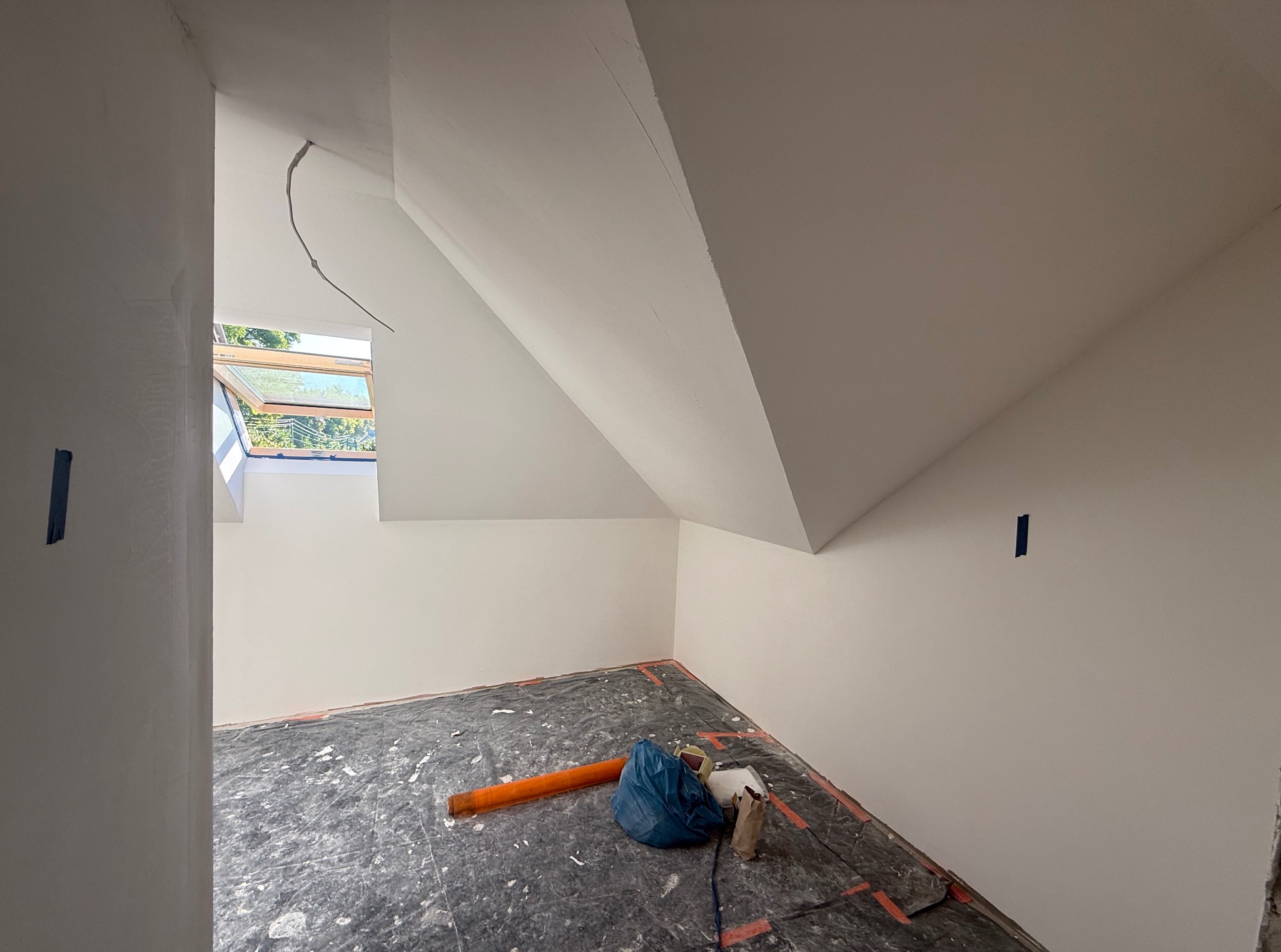 Poddasze po malowaniu na biało, zabezpieczona podłoga folią malarską. Widoczne okno dachowe i fragment instalacji elektrycznej. Świeżo pomalowane ściany i sufit.