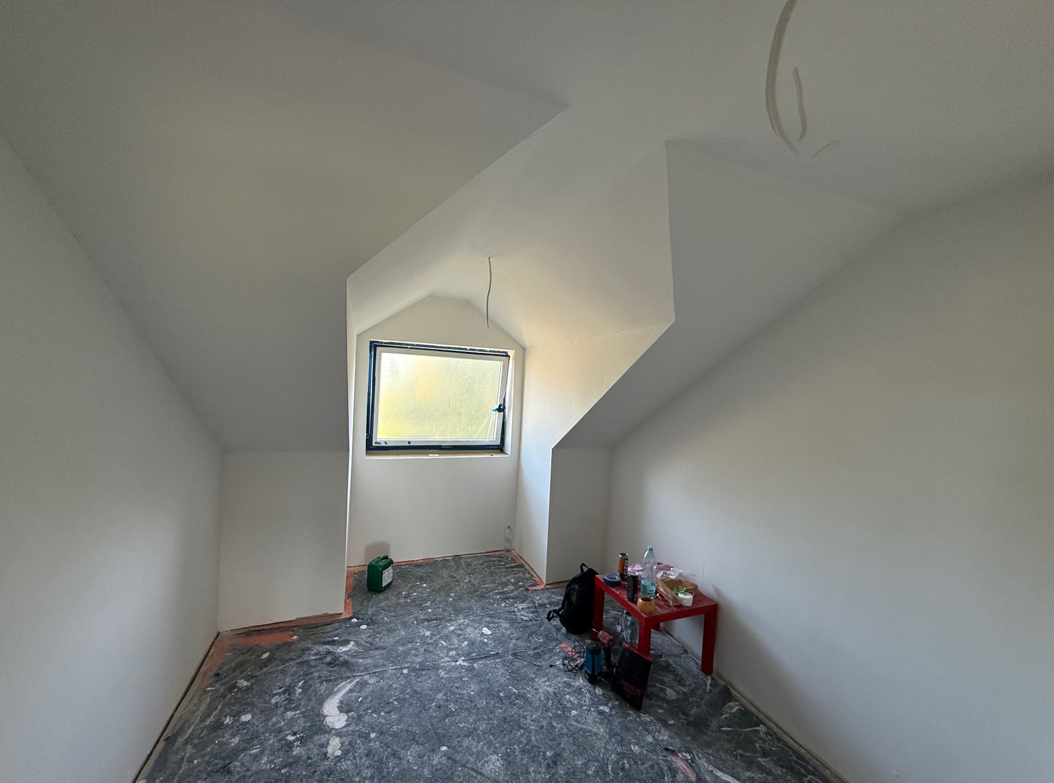 Poddasze po malowaniu na biało, widoczne okno w szczycie, zabezpieczona podłoga, narzędzia i stół z akcesoriami malarskimi. Świeżo pomalowane ściany i sufit.