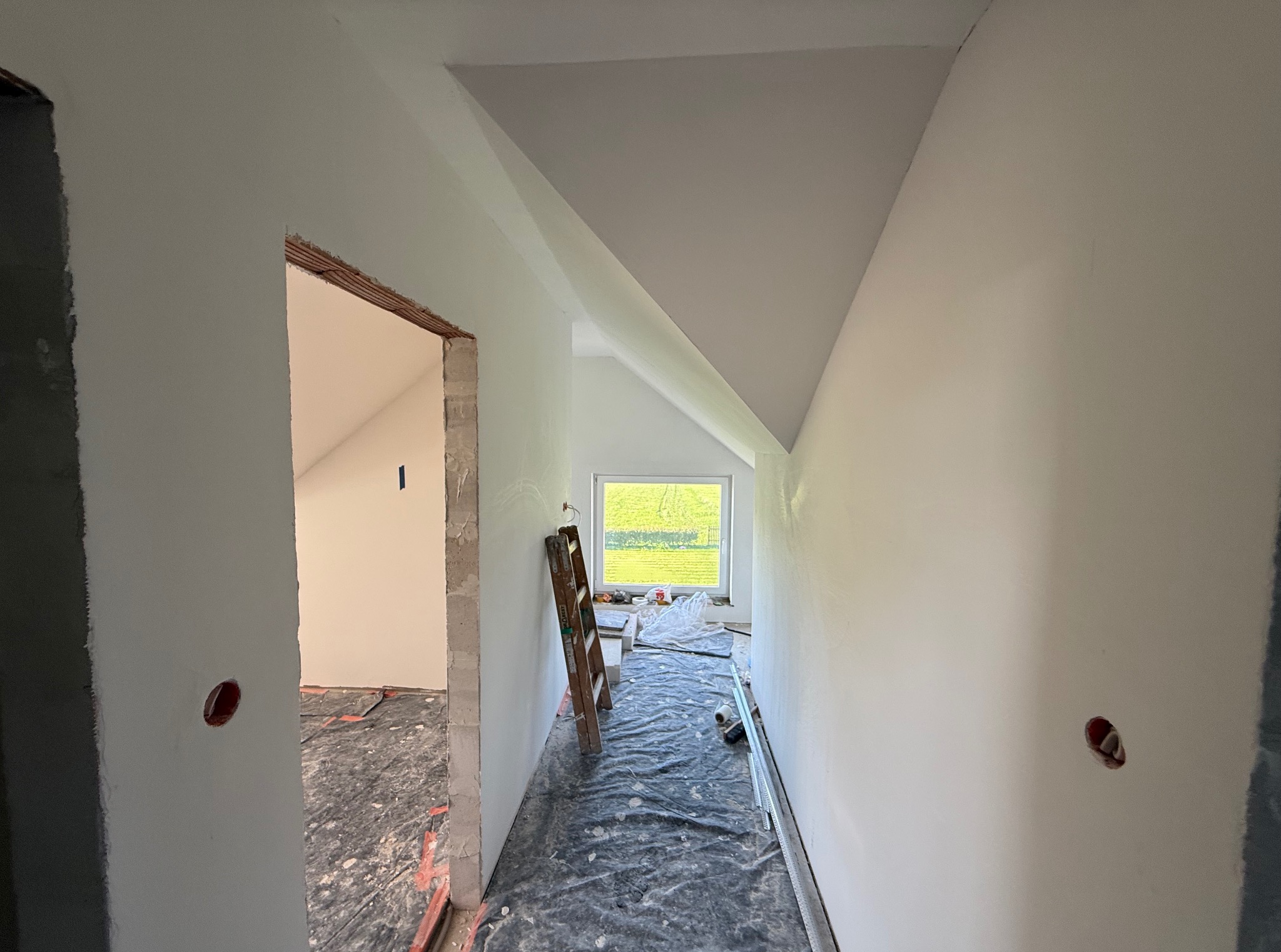 Białe ściany poddasza po malowaniu, widok z korytarza. Widoczne okno, drabina oparta o ścianę i zabezpieczona podłoga. Otwór w ścianie z instalacją elektryczną.