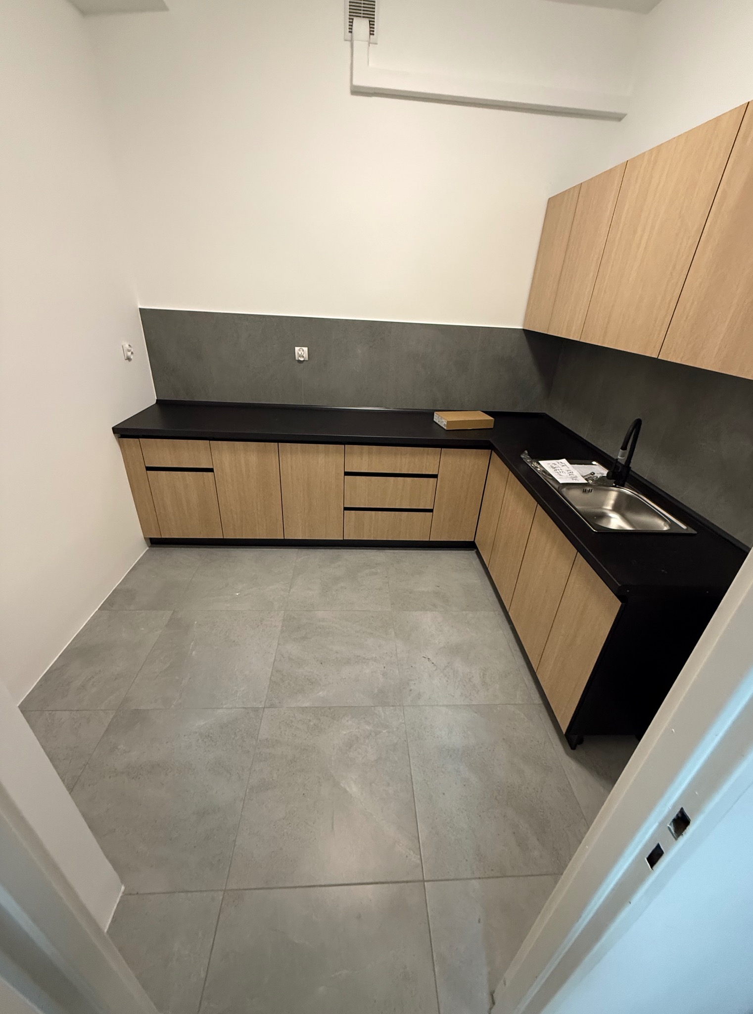 Nowoczesna kuchnia z drewnianymi szafkami i czarnym blatem, szare płytki na podłodze i ścianie. Ujęcie z perspektywy wejścia, widoczny fragment ościeżnicy.