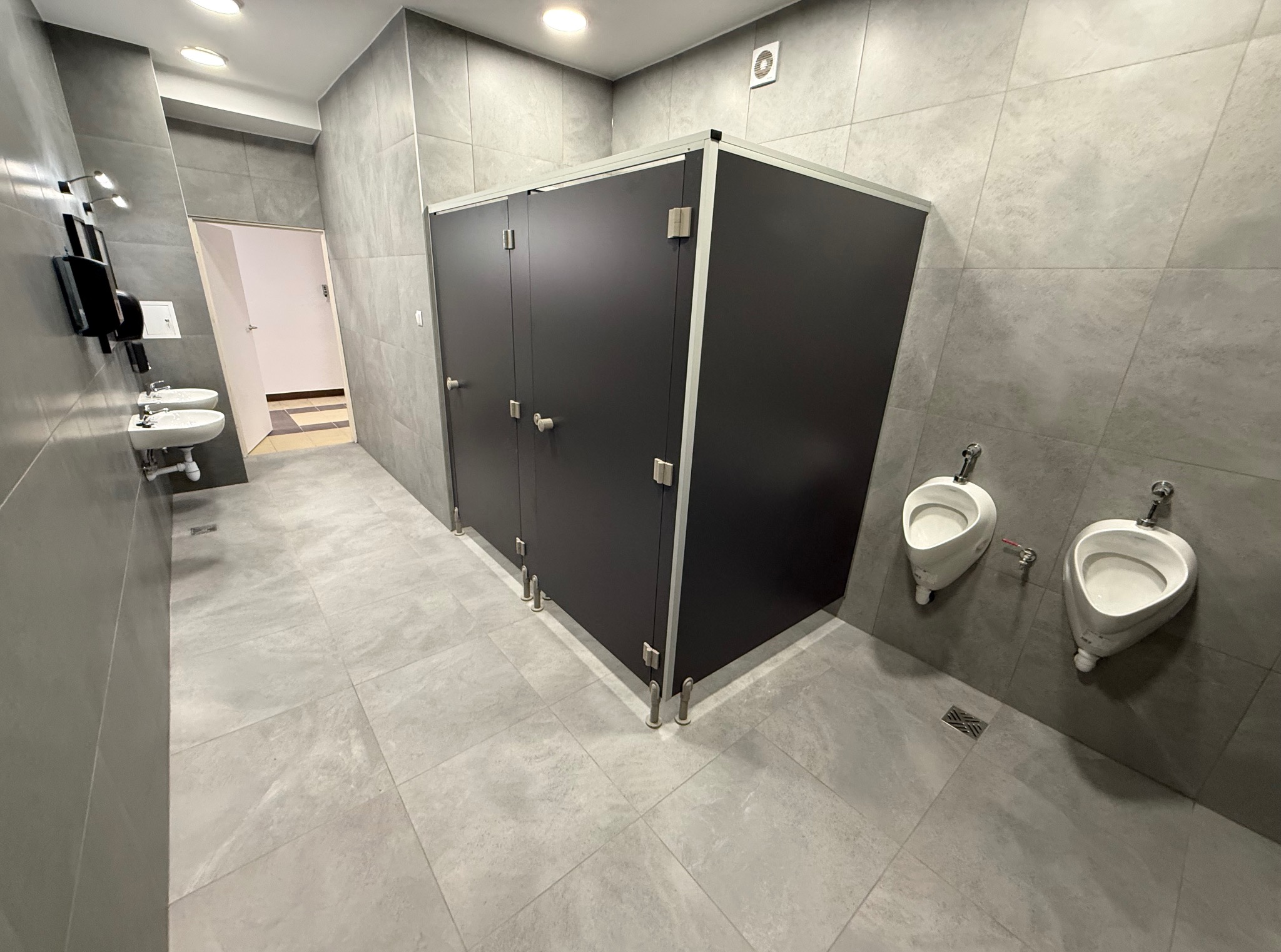 Nowoczesna toaleta publiczna z szarymi płytkami, kabinami WC z ciemnymi drzwiami i białymi pisuarami. Widoczne umywalki w tle. Minimalistyczny design, czyste linie.