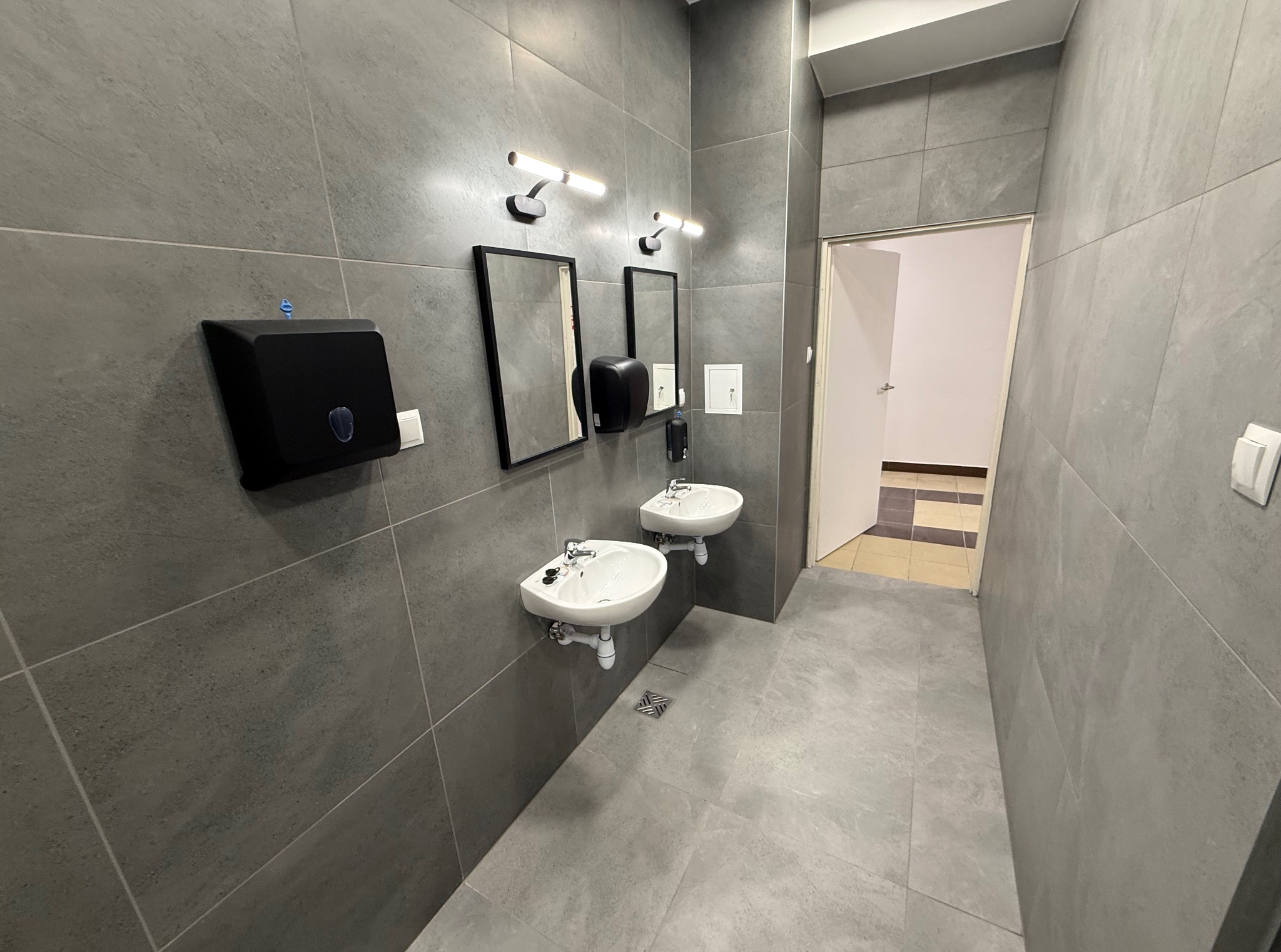 Nowoczesna łazienka publiczna z szarymi płytkami, dwoma umywalkami, lustrami i oświetleniem LED. Widoczny dozownik mydła i podajnik ręczników. Minimalistyczny design.
