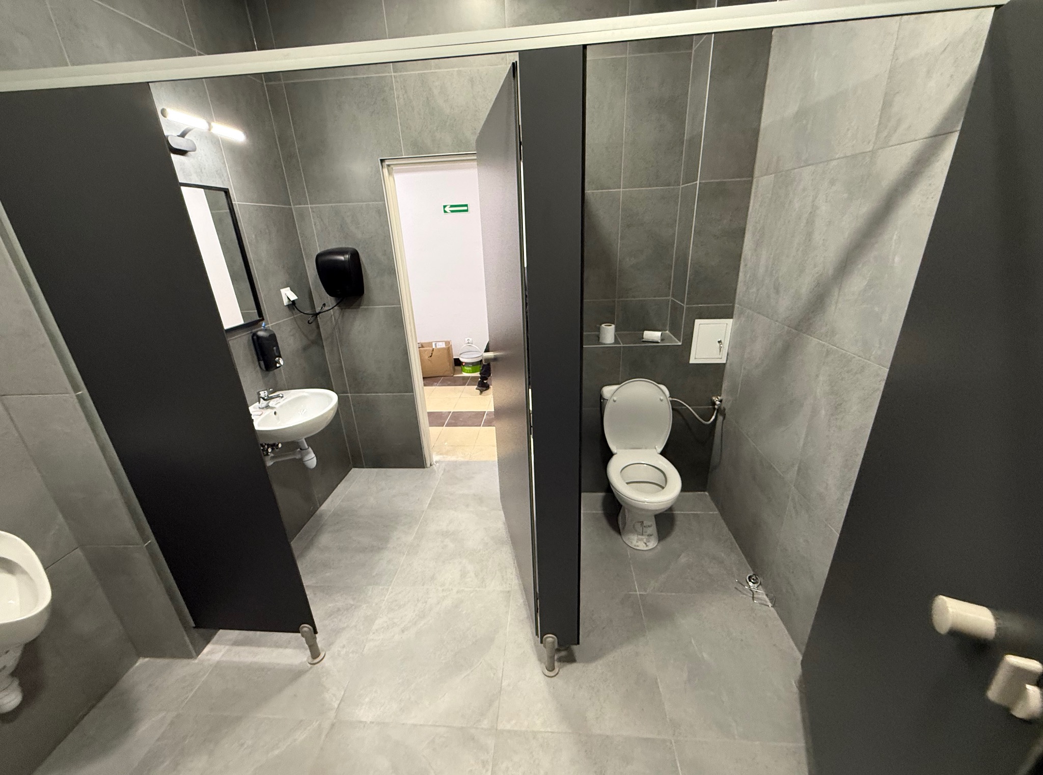 Wnętrze publicznej toalety z szarymi płytkami, kabinami z czarnymi drzwiami i białymi urządzeniami sanitarnymi. Widoczny fragment umywalki i toaleta.