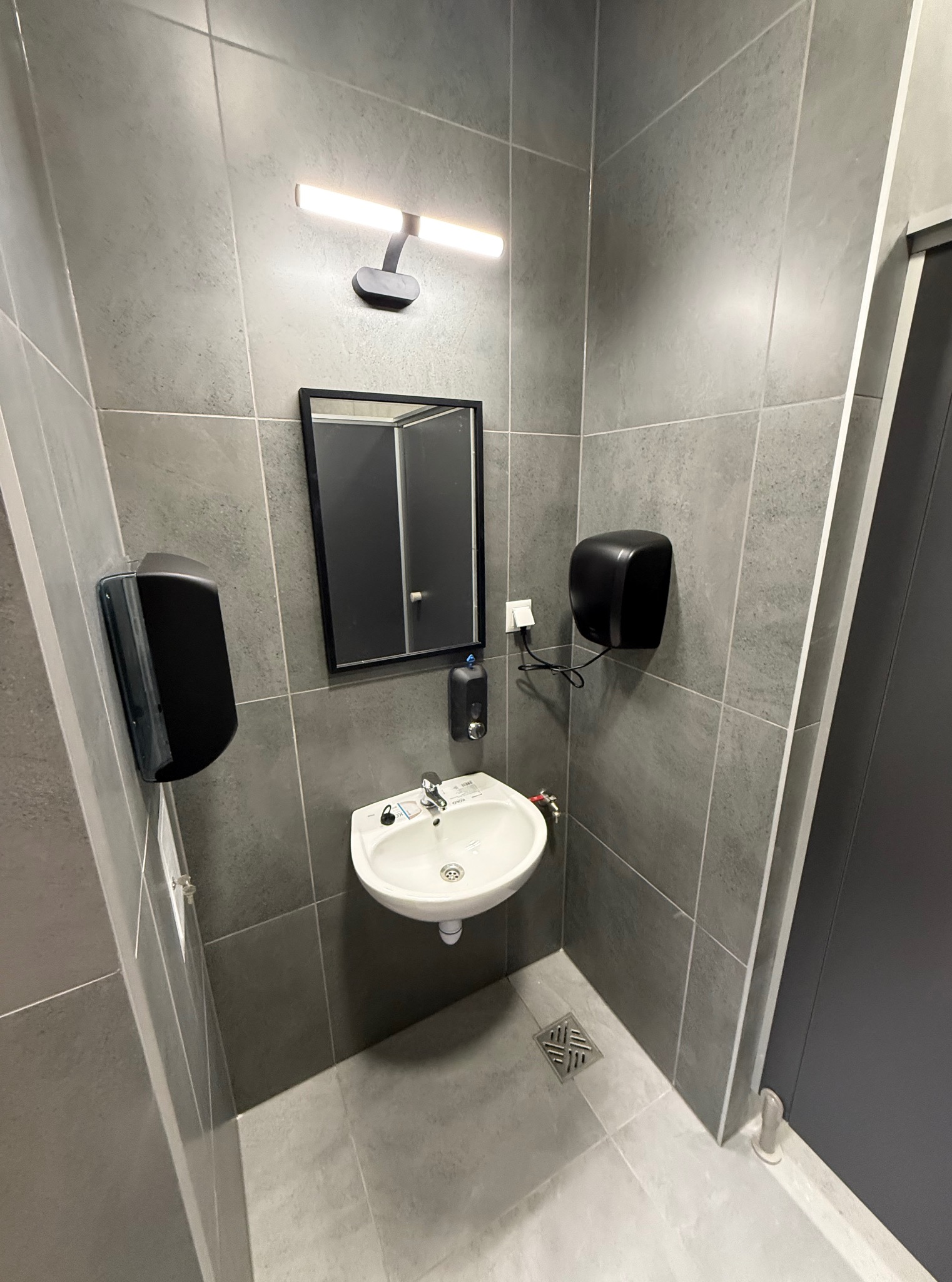 Nowoczesna, szara łazienka z umywalką, lustrem i dozownikiem mydła. Minimalistyczny design, płytki imitujące beton, oświetlenie LED. Widoczny odpływ liniowy.