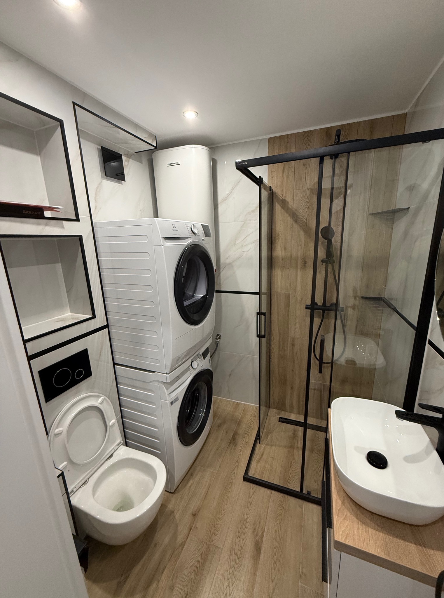 Nowoczesna łazienka po remoncie: kabina prysznicowa typu walk-in z czarnymi ramami, umywalka nablatowa, toaleta podwieszana i pralko-suszarka w zabudowie.