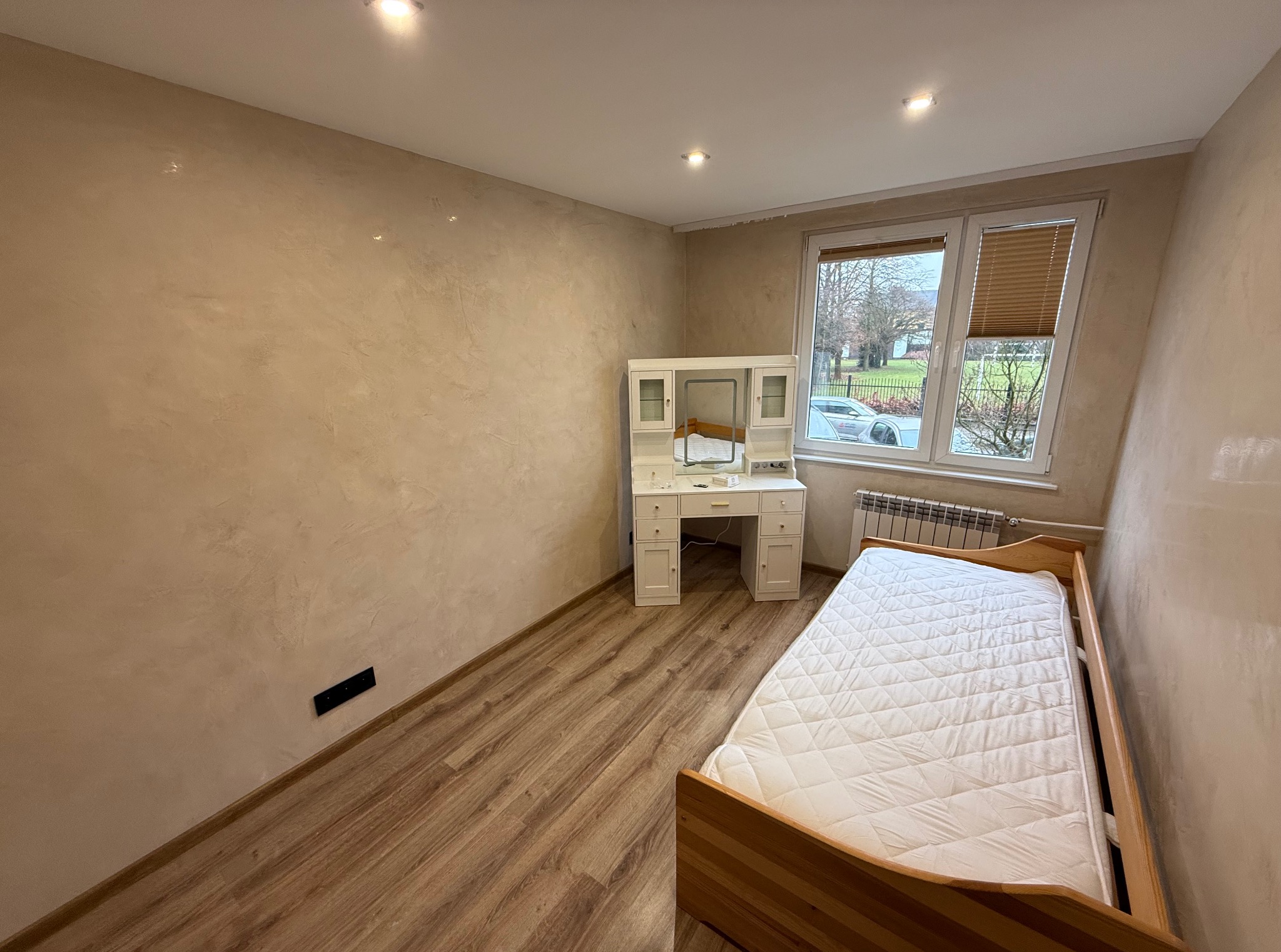 Pokój po odświeżeniu: beżowe ściany z efektem dekoracyjnym, drewniana podłoga, biała toaletka i łóżko. Widok na okno z żaluzjami.