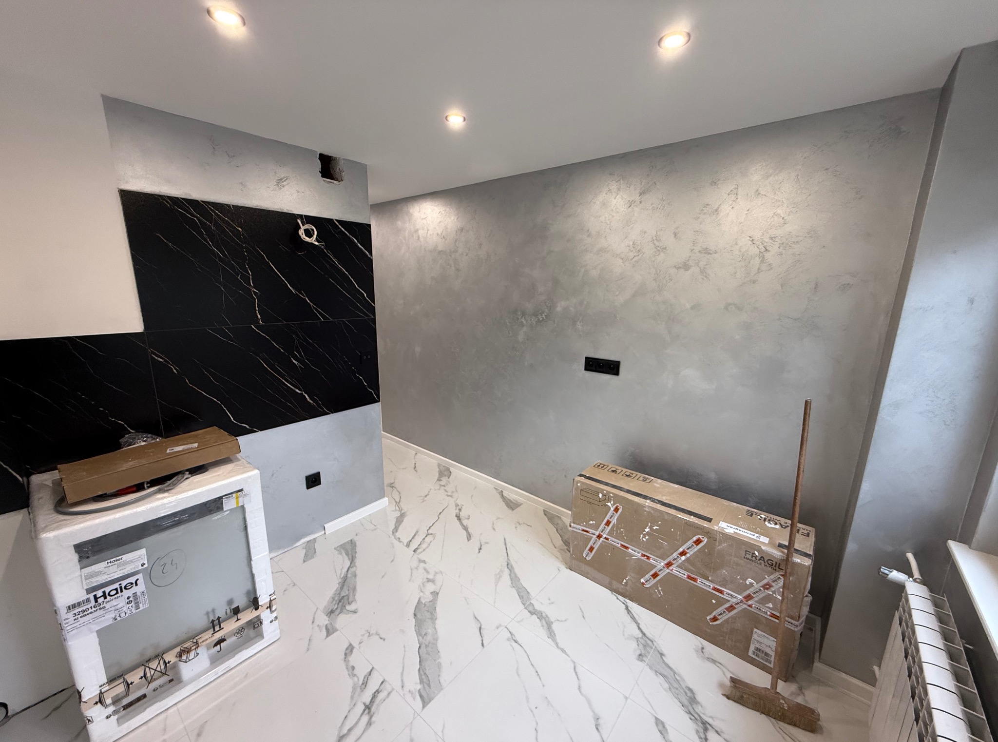 Wykończona kuchnia z marmurową podłogą i ścianą w odcieniach szarości. Widoczna zabudowa z czarnymi płytkami imitującymi marmur oraz sprzęt AGD w opakowaniu.