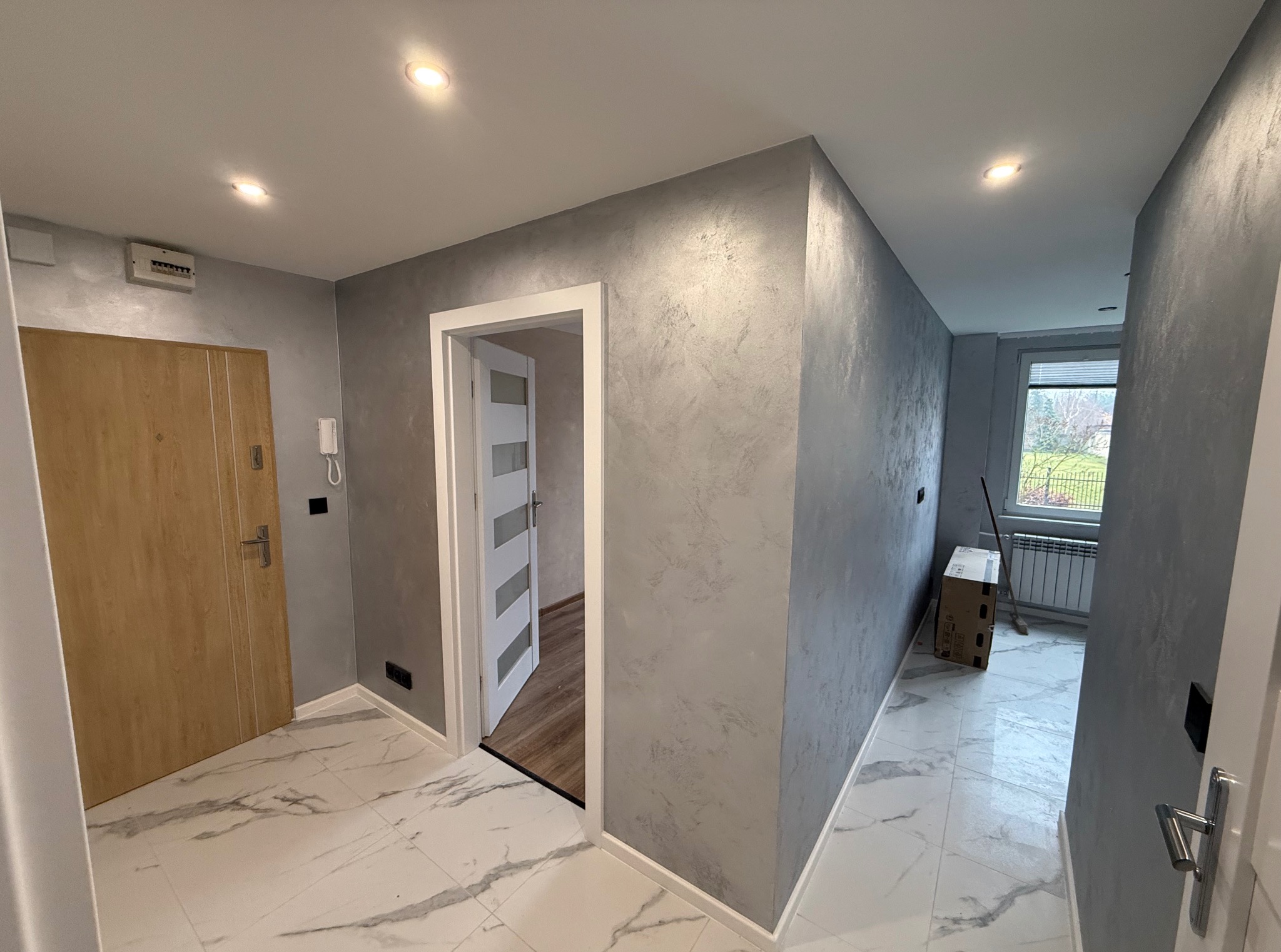 Korytarz po remoncie: szare ściany z efektem dekoracyjnym, biała marmurowa podłoga, drewniane drzwi wejściowe i białe drzwi wewnętrzne. Widoczne oświetlenie sufitowe.