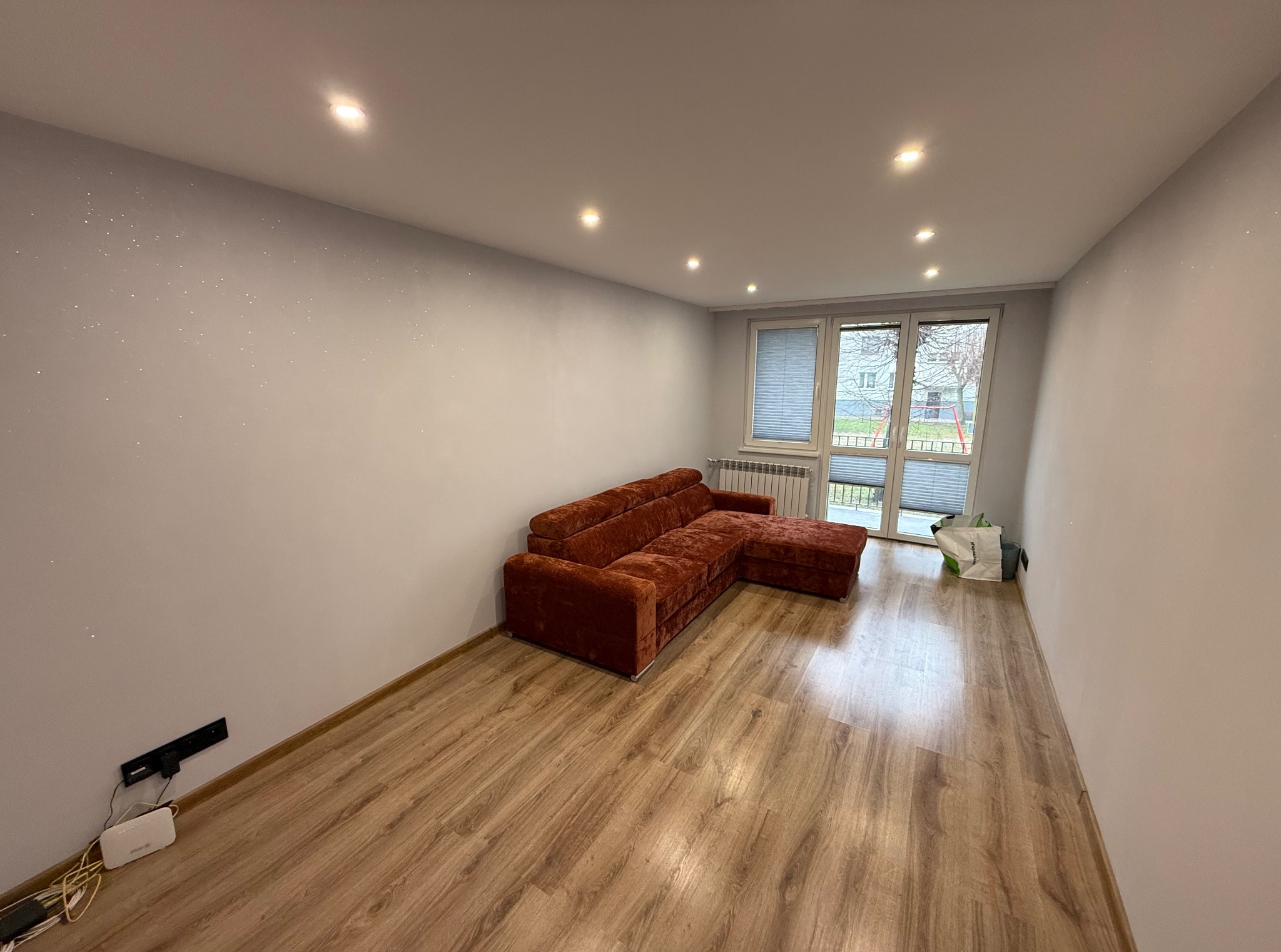 Pokój po remoncie: parkiet, ściany z delikatnym brokatem, brązowa sofa narożna, okno z żaluzjami. Widoczne punkty świetlne w suficie.